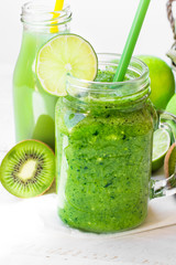 Close up mason jar mug with green fresh vegetable smoothie, bottle with fruit juice, lime slice, kiwi, apples, outdoors, bright sunlight spot, summer, spring