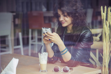 Brunette beautiful stylish caucasian woman in casual outfit on a walk on european city street sitting at the cafe typing message on her phone