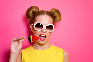 Portrait of stylish cute young blonde woman in sunglasses with colorful watermelon lollipop on pink background - obrazy, fototapety, plakaty