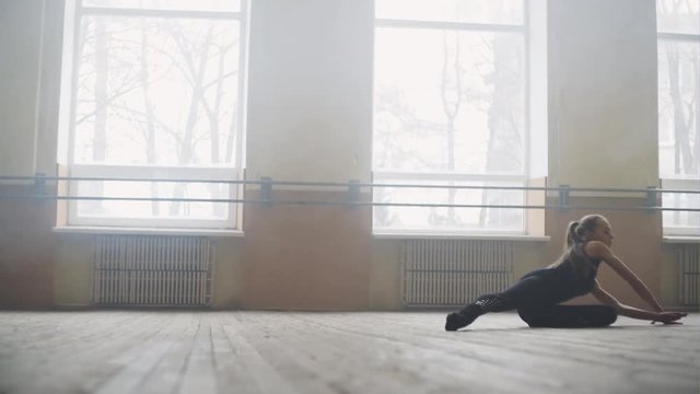 Young girl ballet dancer in black sportswear practicing alone in large empty hall, gracefully doing complex acrobatic moves on the dance floor