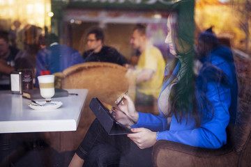 Brunette woman in business clothes: grey sweater and blue jacket sitting at the cafe near the window in european city drinking latte coffee and working using her tablet and smartphone. copy space