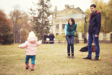 Fototapeta premium Happy loving family(mother, father and little daughter kid) outdoors walking having fun on a park in autumn season. Fallen yellow leaves on a background. Cold weather