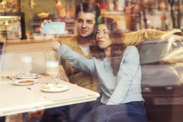 Cute brunette loving couple in cozy warm sweaters on a date at the cafe making selfie on the phone. cold autumn, winter or spring day, european city. indoors