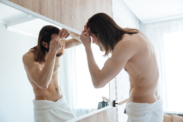 Naked bearded young man in towel squeezing pimple near mirror
