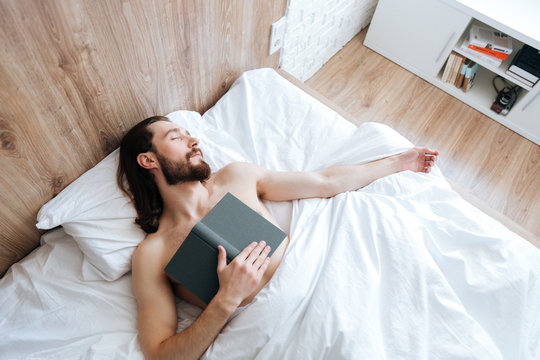 Tired bearded young man with book sleeping in bed
