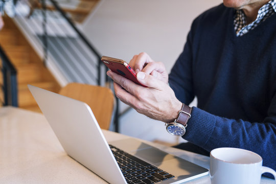 Midsection Of Man Using Smart Phone While Sitting By Laptop Computer At Table