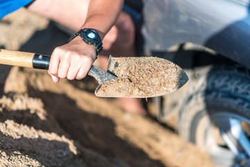 a man digs a car stuck in the sand, throwing sand shovel out from under the car