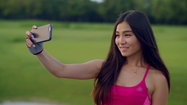 Smile girl taking selfie outdoors. Close up of sporty woman taking selfie photo on smartphone in park. Asian girl posing for selfie with phone. Happy girl posing for selfie portrait. Fit girl selfi