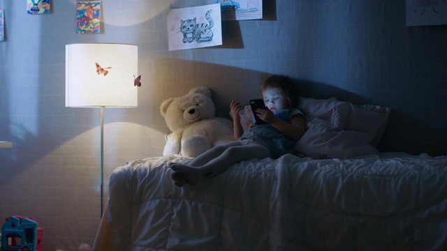 Cute Little Girl Lies on Her Bed and Watches Cartoons on a Smartphone. Her Floor Lamp is On. Shot on RED EPIC-W 8K Helium Cinema Camera.