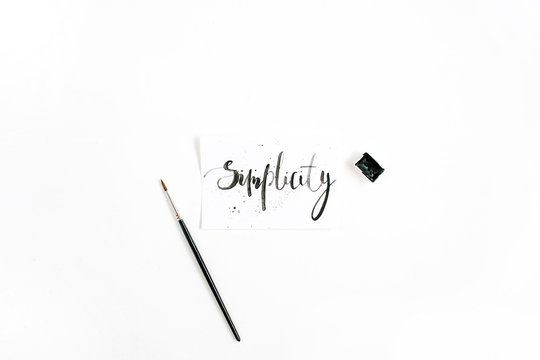 Minimalistic stylish composition with word Simplicity written in calligraphic style on paper with paint brush on white background. Flat lay, top view
