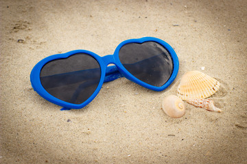 Fototapeta na wymiar Blue sunglasses shaped heart with shells on the sand