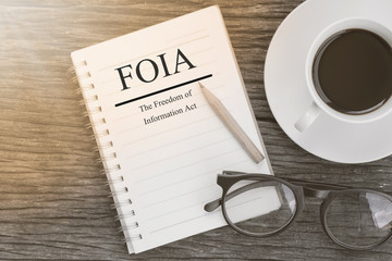 Concept  FOIA (The Freedom of Information Act) message on notebook with glasses, pencil and coffee cup on wooden table.