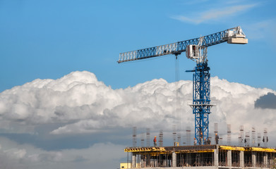 The construction tower crane is blue next to the builders on top of a building under construction. On a background of clouds and blue sky