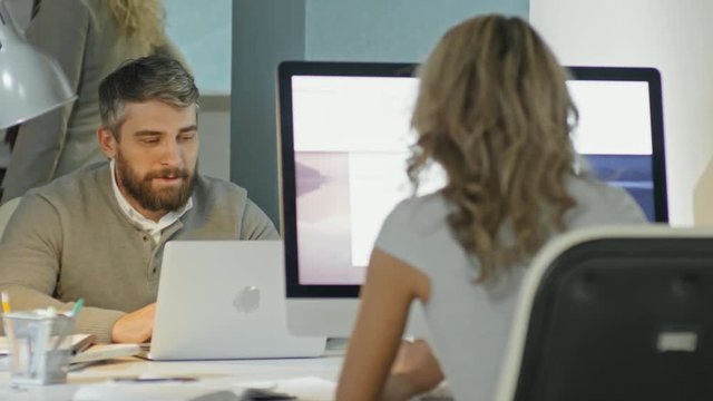 PAN of businessman with beard working on laptop and discussing work with female colleague sitting opposite him 