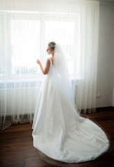 Fototapeta na wymiar Bride in long white dress with long veil stands before bright window on wooden floor