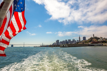 San Francisco Financial District cityscape and Oakland Bridge on sunny day, California, United States. Sea views from Alcatraz boat with American flag waving. Freedom and travel concept.