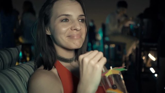 Young, happy woman drinking cocktail sitting on terrace in bar at night
