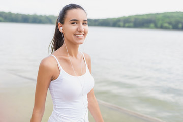 Young beautiful brunette smiling woman listening to music at park and lake. Close up face of smiling woman with earphone looking at camera. Beautiful girl running at park and listening to music.