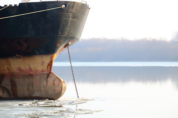 Prow of old rusty dark ship with anchor chain anchored in winter frozen Danube river