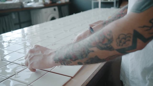 Tattooed hands precisely apply white ceramic tile on wooden kitchen table next to other tiles, separated with plastic spacers. DIY renovation process