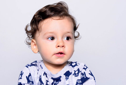 Portrait of a cute curly hair baby boy looking away. Adorable one year old child looking funny and curious to the side. Baby boy with big eyes and open mouth showing his teeth.