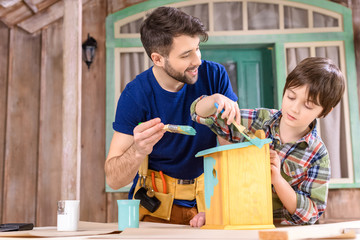 Happy father and son painting small wooden birdhouse on porch