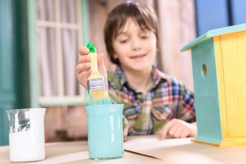 Cute smiling boy holding paintbrush while painting handmade birdhouse