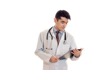 young serious brunette man doctor in uniform with stethoscope on his shoulders make notes isolated on white background