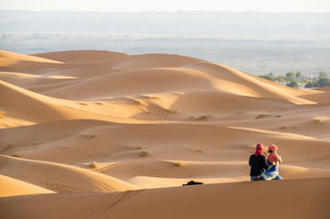 Fototapeta na wymiar Two girls wearing a turban are sitting on a sand dune admiring the sunset in Merzouga, Morocco.