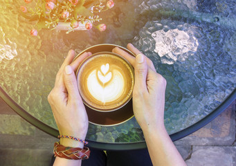Hands with bracelets on wrist  holding cup of hot coffee latte cappuccino on round glass top table, filtered color effect.