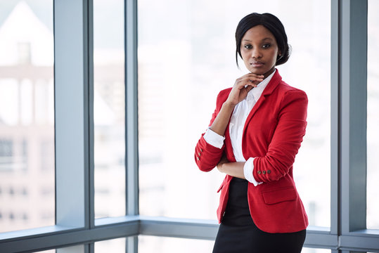 Confident businesswoman looking at the camera with bold body language while wearing a red blazer with large windows behind her in the background.