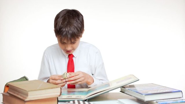 Young school kid with red tie sitting at table with books and atlas playing with cube on white background in slowmotion