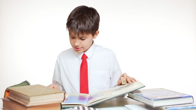 Young concentrated school kid with red tie studying atlas sitting at table with books on white background in slowmotion