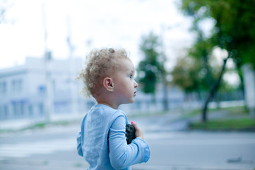 Little beautiful girl with short curly hair, walks around the city. The child holds a toy and is near the road.