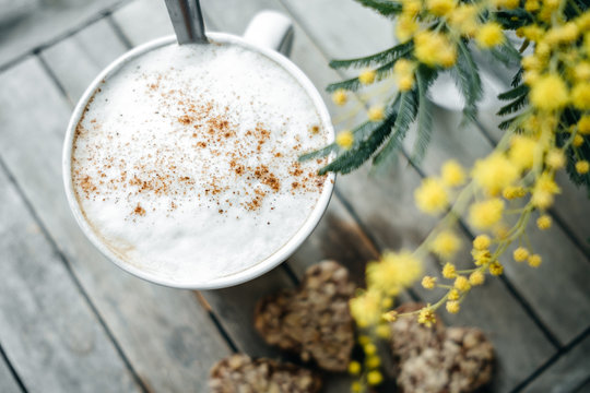 Top view of Hot cocoa with cinnamon in  white mug with heart shape cookies and mimoza on wooden table. Spring concept