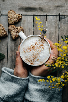 Womans hand with coffee with cinnamon in  white mug with heart shape cookies and mimoza on wooden table. Spring concept