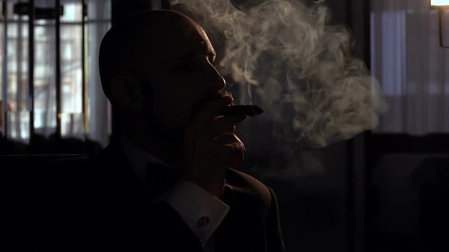 Adult man smokes a cigar in the dark with artificial light, slow motion.