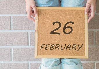 Closeup calendar at the cork board in hand of asian woman in front of her legs with 26 february word on brick wall textured background