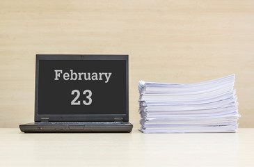 Closeup computer laptop with february 23 word on the center of screen in calendar concept and pile of work paper on wood desk and wood wall in work room textured background with copy space