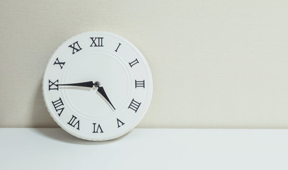 Closeup white clock for decorate show a quarter to five p.m. or 4:45 p.m. on white wood desk and cream wallpaper textured background with copy space