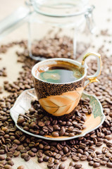 Cup of coffee surrounded by a scattered coffee beans from a jar of glass on a wooden table - vertical view