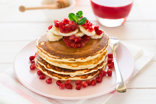 Traditional breakfast: stack of pancakes with banana slices and garnet seeds on white wooden table. Selective focus