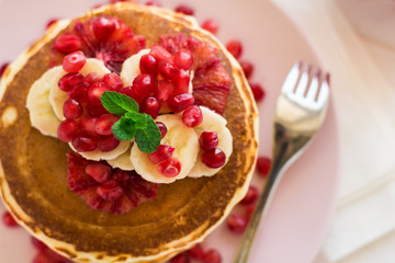 Traditional breakfast: stack of pancakes with banana slices and garnet seeds on white wooden table. Selective focus