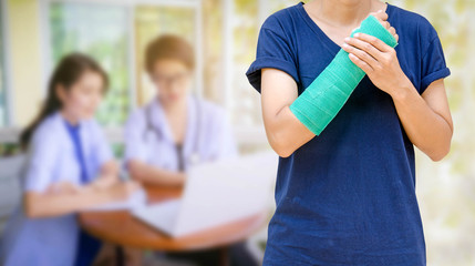 Green cast on an arm of a women on blurred two female doctor with stethoscope, clipboard and Laptop on desk,Doctor working in clinic writing a prescription,  Health care and medical concept