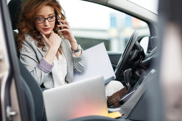 Beautiful female sales manager using laptop and phone in the car.Preparing and looking into contract papers.
