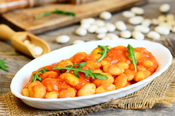 Spicy baked beans with tomato sauce served with fresh arugula on a plate. Slow cooked home baked beans recipe. Vegetarian side dish. Rustic style. Closeup