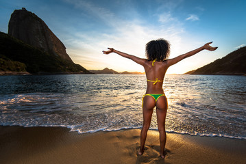 Fototapeta na wymiar Sexy Brazilian Girl in Bikini Stands in the Beach With Open Arms and Welcomes A New Day in Rio de Janeiro With the Sugarloaf Mountain in the Background