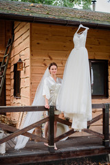 Pretty bride in silk robe stands behind her wedding dress on wooden porch