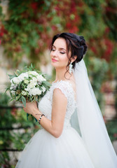 Gorgeous bride in luxury dress poses before wall with green and red ivy