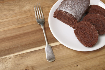 A sliced chocolate Swiss roll cake on a wooden counter top background, with fork and blank space at side